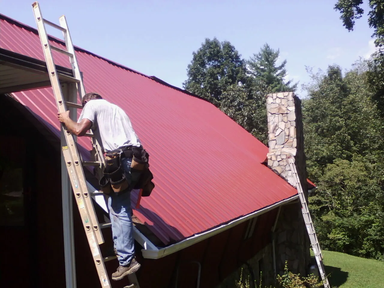 Roofer on a ladder beside a red metal roof and stone chimney — Soffit & Fascia Repair by Roofing Brothers in Drexel Hill